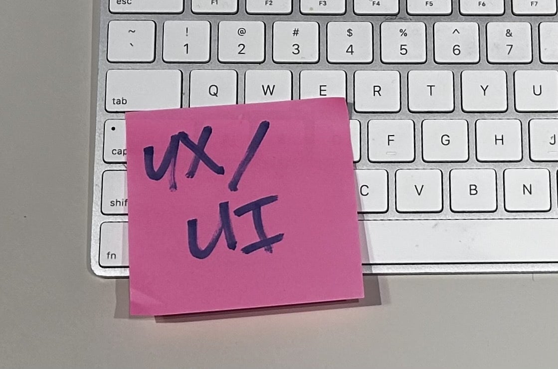 A pink Post-It note with "UX/UI" written in Sharpie, stuck on a Mac keyboard